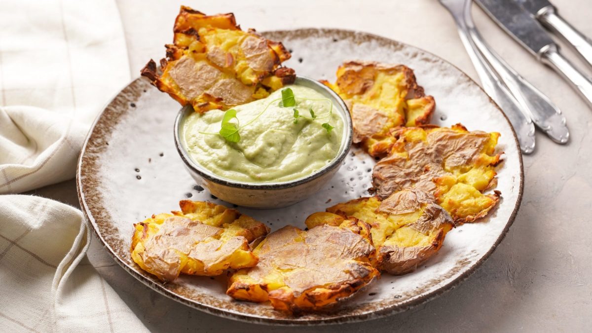 Ein grauer Teller mit den Quetschkartoffeln auf beigem Untergrund. In der Mitte ein Schälchen mit der hellgrünen Mayonnaise und oben rechts im Bild silbernes Besteck.