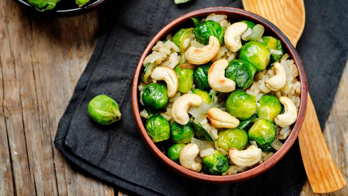 Draufsicht: eine Rosenkohl-Cashew-Pfanne mit Reis auf einem Filz, der auf einer Holzplatte liegt. Daneben liegt ein Holzlöffel.