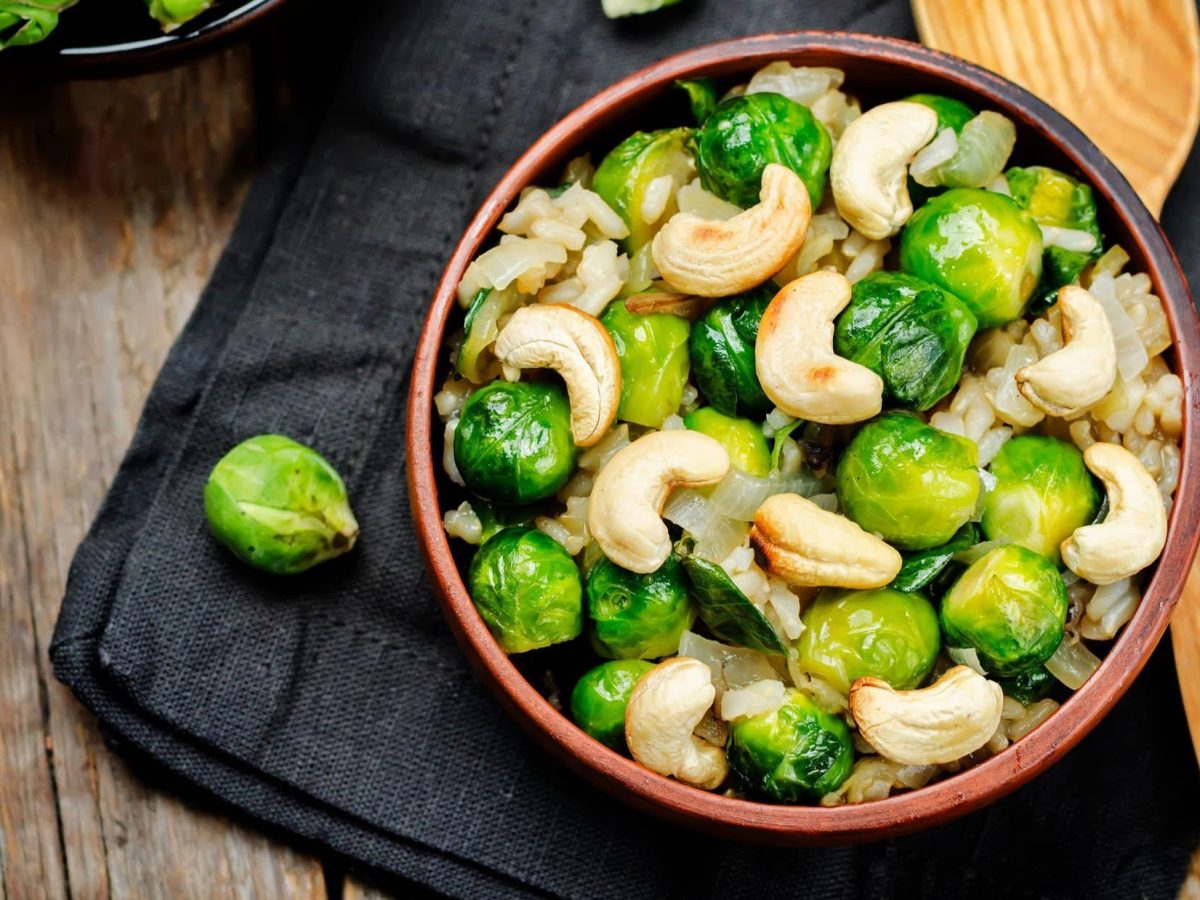 Draufsicht: eine Rosenkohl-Cashew-Pfanne mit Reis auf einem Filz, der auf einer Holzplatte liegt. Daneben liegt ein Holzlöffel.