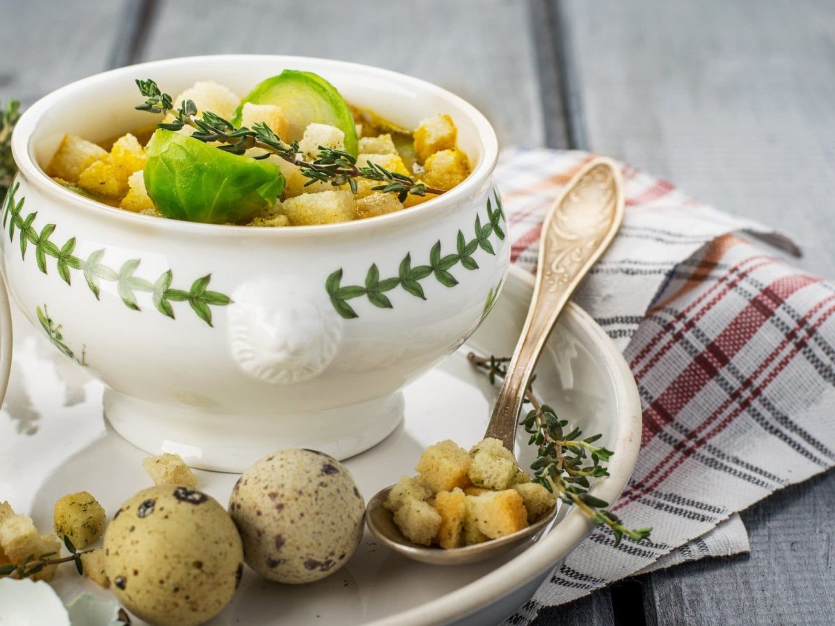 Frontal: Eine kleine Suppenschüssel mit einer Rosenkohl-Croûton-Suppe mit Wachtelei steht auf einem Teller und der auf einem Untergrund aus Holz. Drum herum liegen frischer Rosenkohl und Thymianzweige.