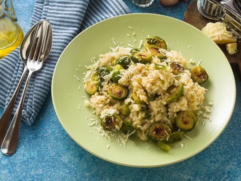 Draufsicht, schräg: Ein grüner Teller mit einem Rosenkohl-Risotto. Drum herum liegen Besteck, Gewürze und andere Zutaten.