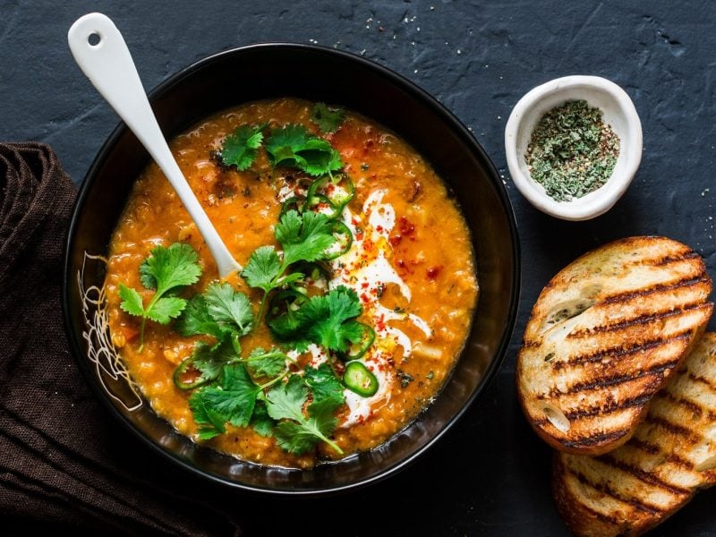 Eine schwarze Schüssel mit der Linsensuppe auf schwarzem Untergrund mit zwei Scheiben geröstetem Weißbrot und einem braunen Tuch. Alles direkt von oben fotografiert.