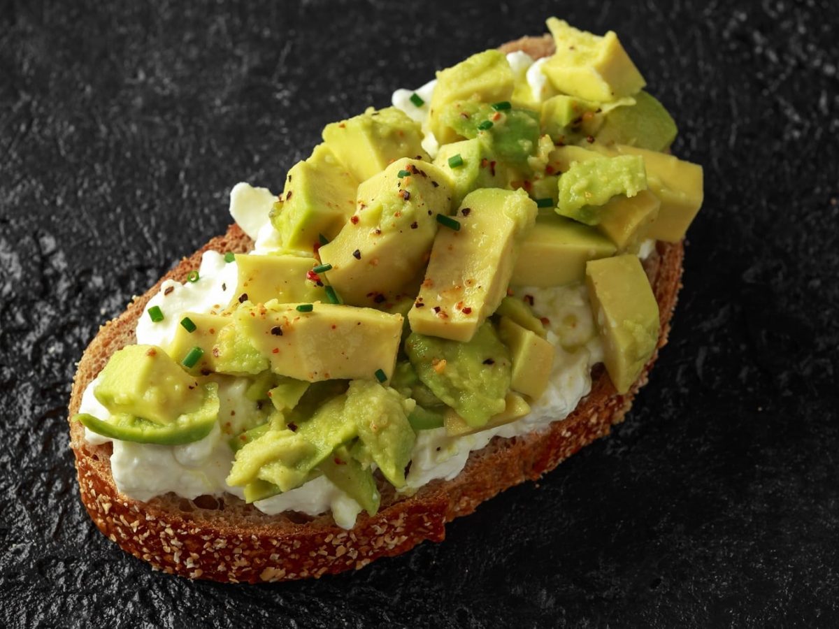 Ein belegtes Brot mit Avocado und körnigem Frischkäse auf dunklem Untergrund von schräg oben nah fotografiert.