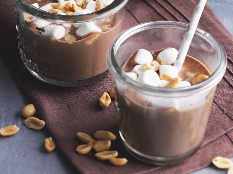 Süß-salzige heiße Schokolade in zwei Gläsern mit Marshmallows und Erdnüssen auf braunem Stoff, grauer Untergrund.