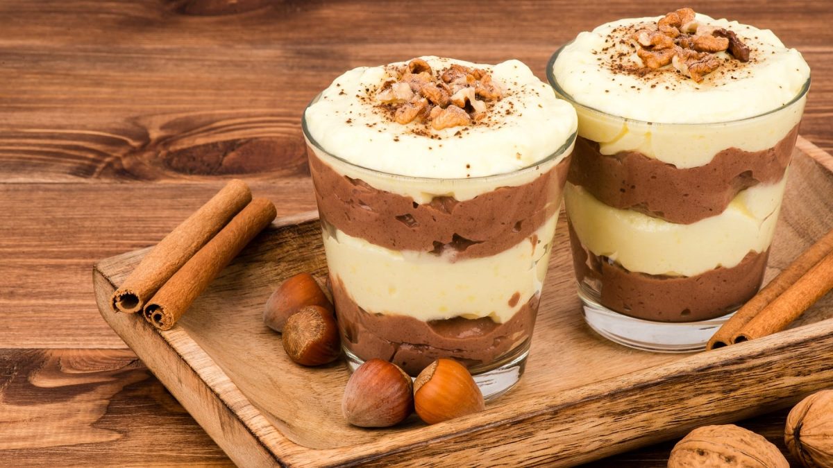 Zwei Gläser mit dem Vanille-Lebkuchen-Schichtdessert im Glas auf einem Holztisch mit Zimt und Nüssen.