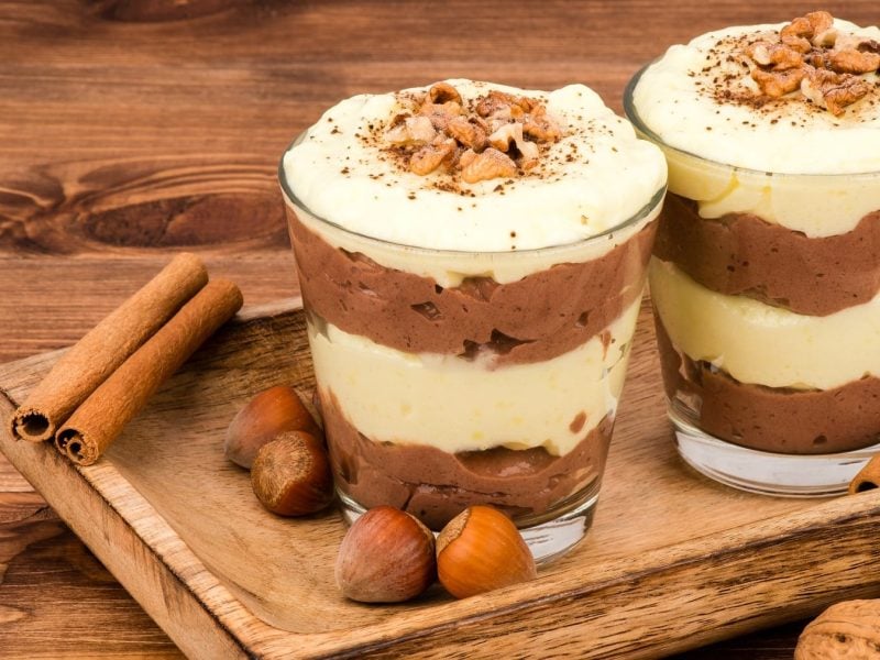 Zwei Gläser mit dem Vanille-Lebkuchen-Schichtdessert im Glas auf einem Holztisch mit Zimt und Nüssen.