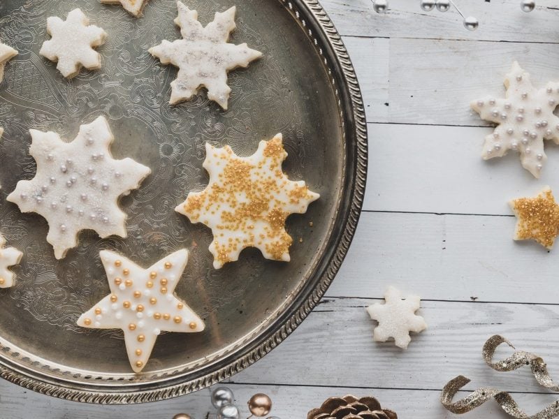 Draufsicht Selbst gebackene Vanillesterne mit einer Schicht weißem Zuckerguss und goldenen und silbernen Deko-Elementen liegen auf einem silbernen Tablett, dass auf einer weißen Holzplatte liegt.