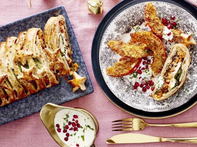 Veggiebraten "Wellington" mit Knusperkürbis auf rosarotem Hintergrund. Rechts davon ein weißer Teller angerichtet mit einer Portion. Unten ein Schälchen mit Dip und rechts davon goldenes Besteck.