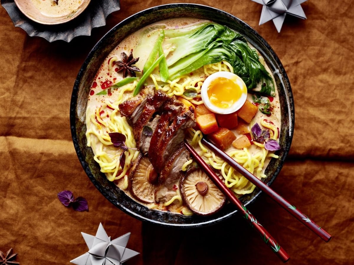 Eine Bowl mit Weihnachtsramen Eat-Club-Style auf hölzernem Hintergrund. Oben ein Teelicht und zwei Geschenkpapierschleifen und Gewürze verteilt auf dem Hintergrund.