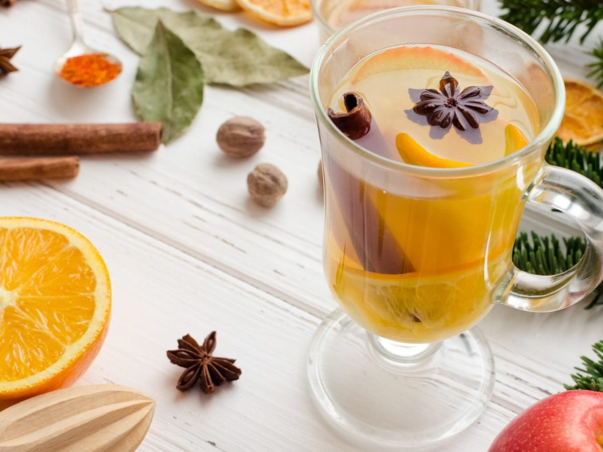 Ein Glas mit dem weißen Glühwein auf einem hellen Holztisch mit allerlei Gewürzen und winterlichen Zutaten.