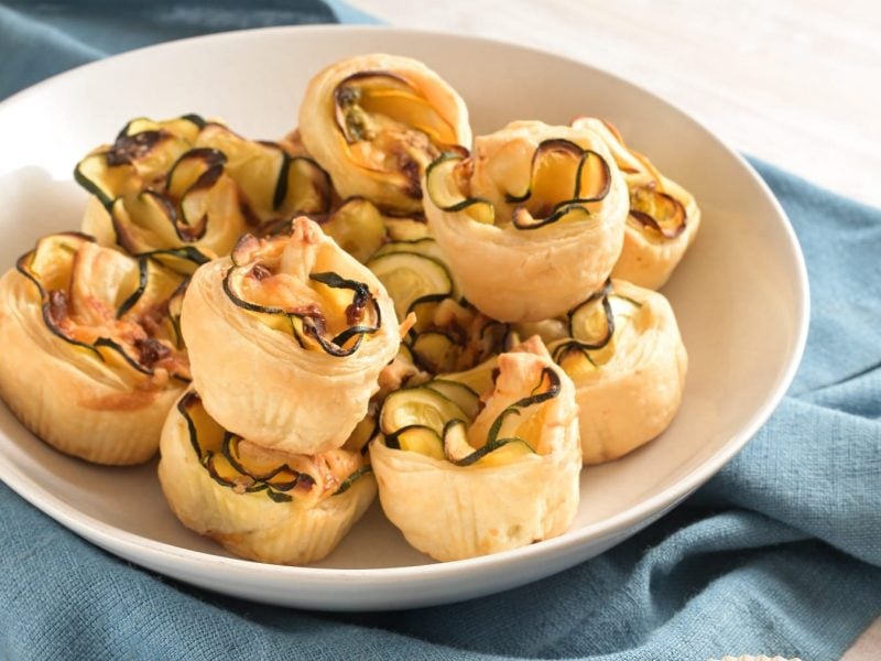 Die Zucchini-Feta-Rosen auf einem weißen Teller und blauem Tuch. Alles schräg von der Seite fotografiert.