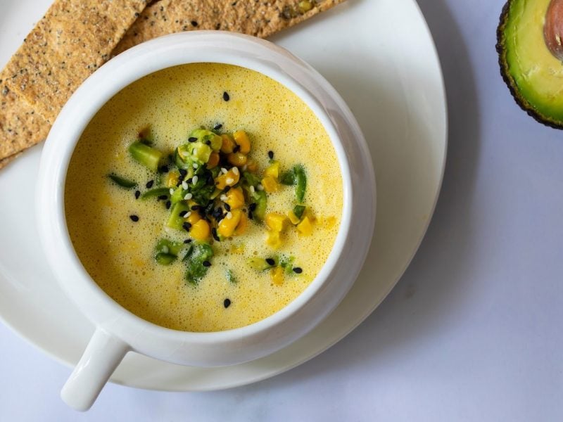 Die Avocado-Maissuppe in hellem Geschirr auf einem grauen Tisch direkt von oben fotografiert.