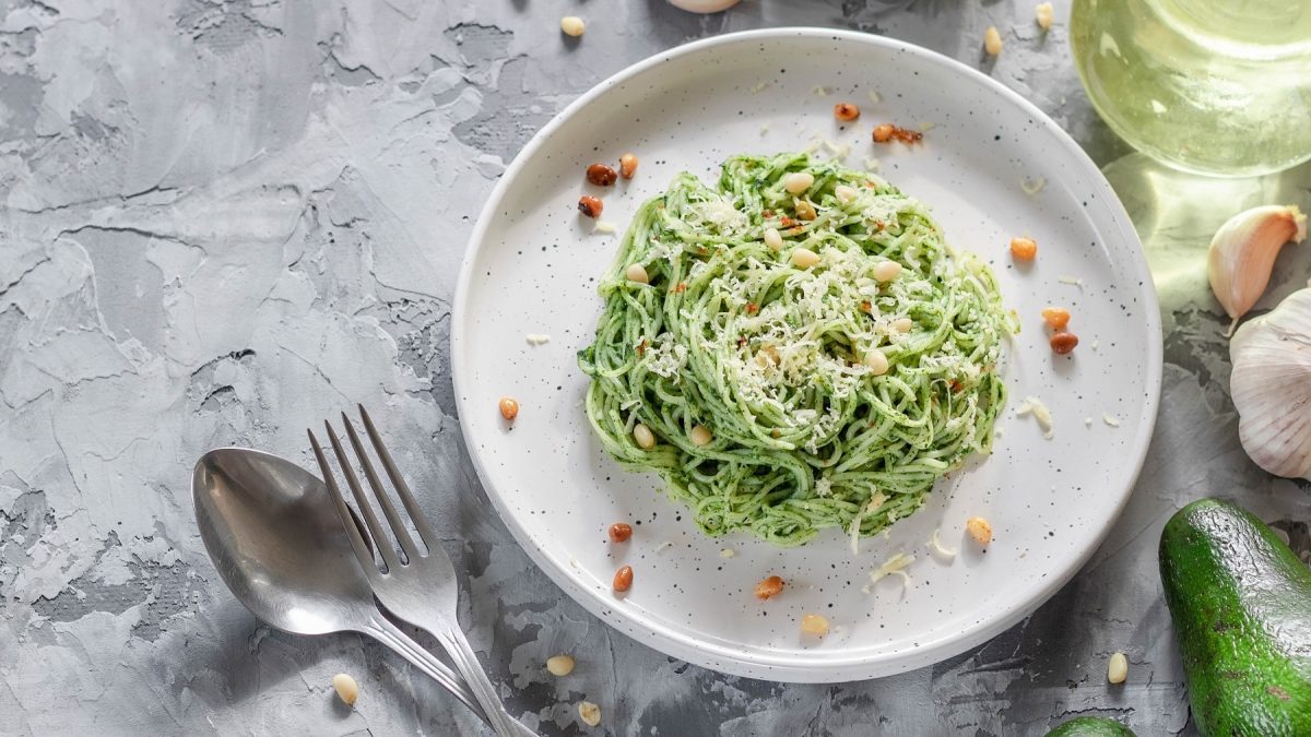 Ein weißer Teller mit der Avocado-Spinat-Pasta auf einem grauen Tisch mit Avocado, Knoblauch und Öl. Alles direkt von oben fotografiert.