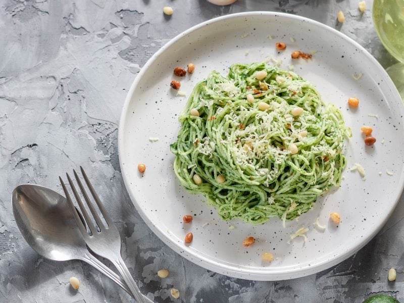 Ein weißer Teller mit der Avocado-Spinat-Pasta auf einem grauen Tisch mit Avocado, Knoblauch und Öl. Alles direkt von oben fotografiert.