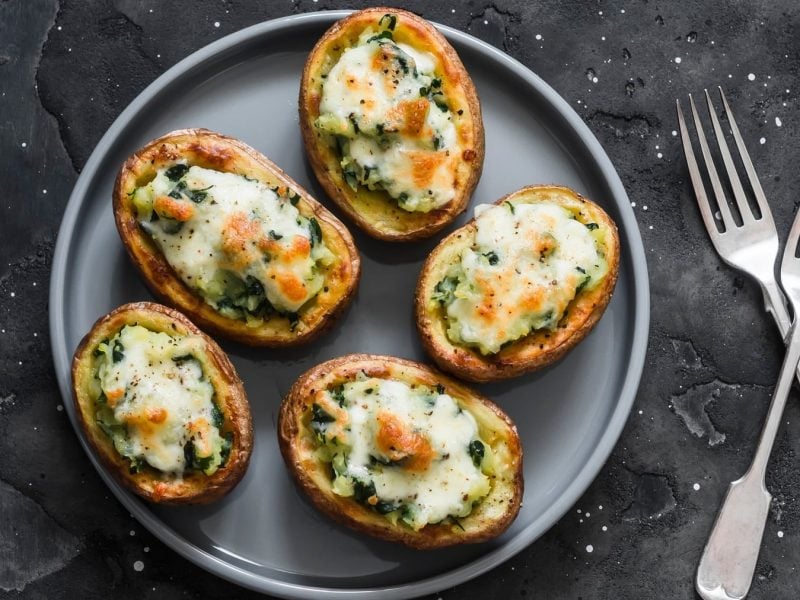 Mehrere Kartoffelhälften mit Spinat und Käse überbacken auf einem grauen Teller und dunklem Untergrund.