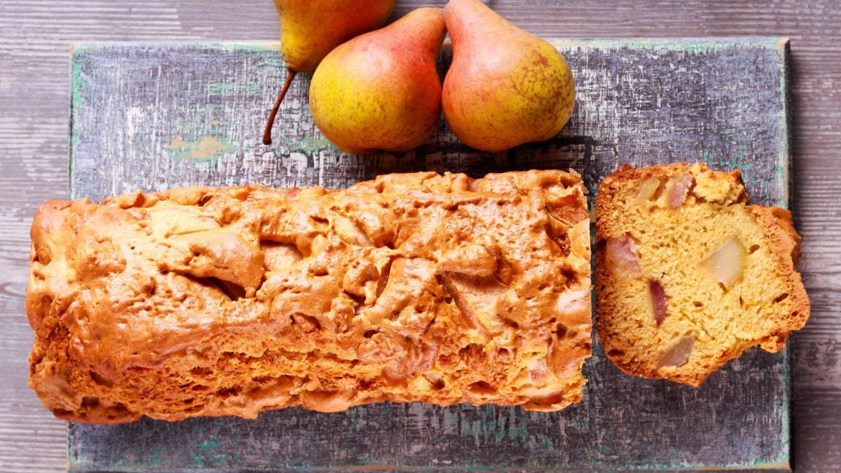 Der angeschnittene Birnen-Kastenkuchen mit einer Scheibe auf grauem Brett. Alles direkt von oben fotografiert.