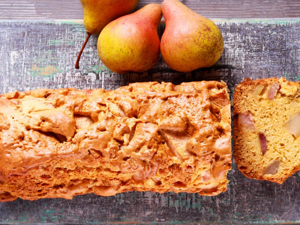 Der angeschnittene Birnen-Kastenkuchen mit einer Scheibe auf grauem Brett. Alles direkt von oben fotografiert.