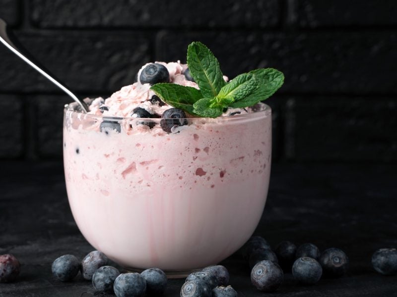 Ein Glas mit der Creme und Minze auf dunklem Untergrund mit Blaubeeren und schwarzem Hintergrund. Alles direkt von der Seite fotografiert.