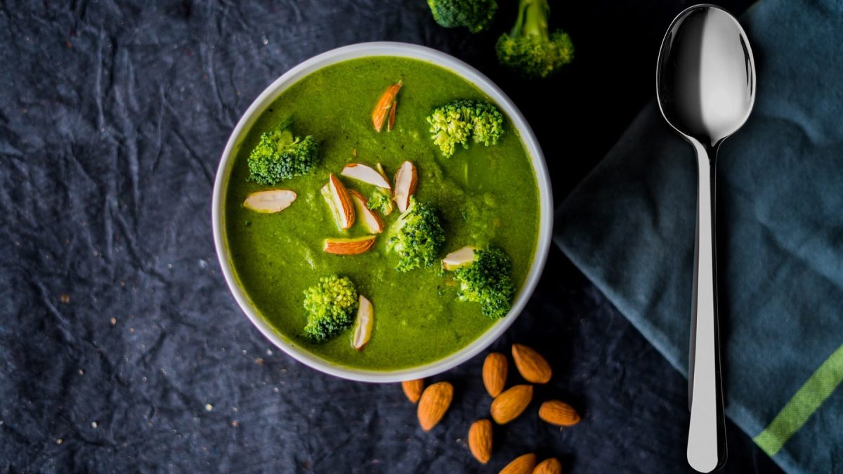 Eine Schüssel mit der Suppe auf dunklem Untergrund. Daneben ein Löffel, Mandeln und einige Brokkoli-Röschen. Alles direkt von oben fotografiert.