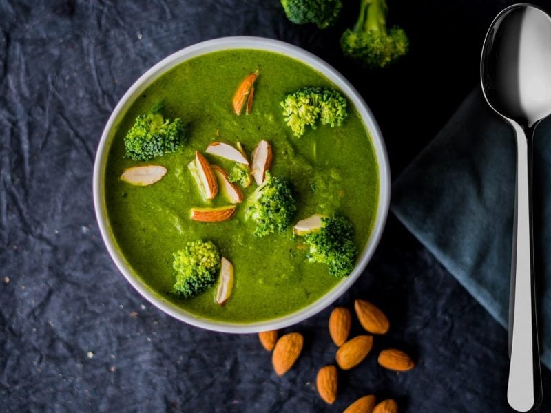 Eine Schüssel mit der Suppe auf dunklem Untergrund. Daneben ein Löffel, Mandeln und einige Brokkoli-Röschen. Alles direkt von oben fotografiert.