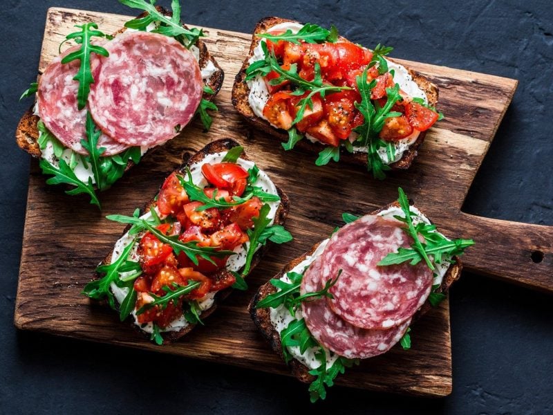 Draufsicht: Dunkler Untergrund, ein Holzbrett mit Bruschetta mit Fenchelsalami. Zwei sind zudem mit Tomatenwürfeln belegt.