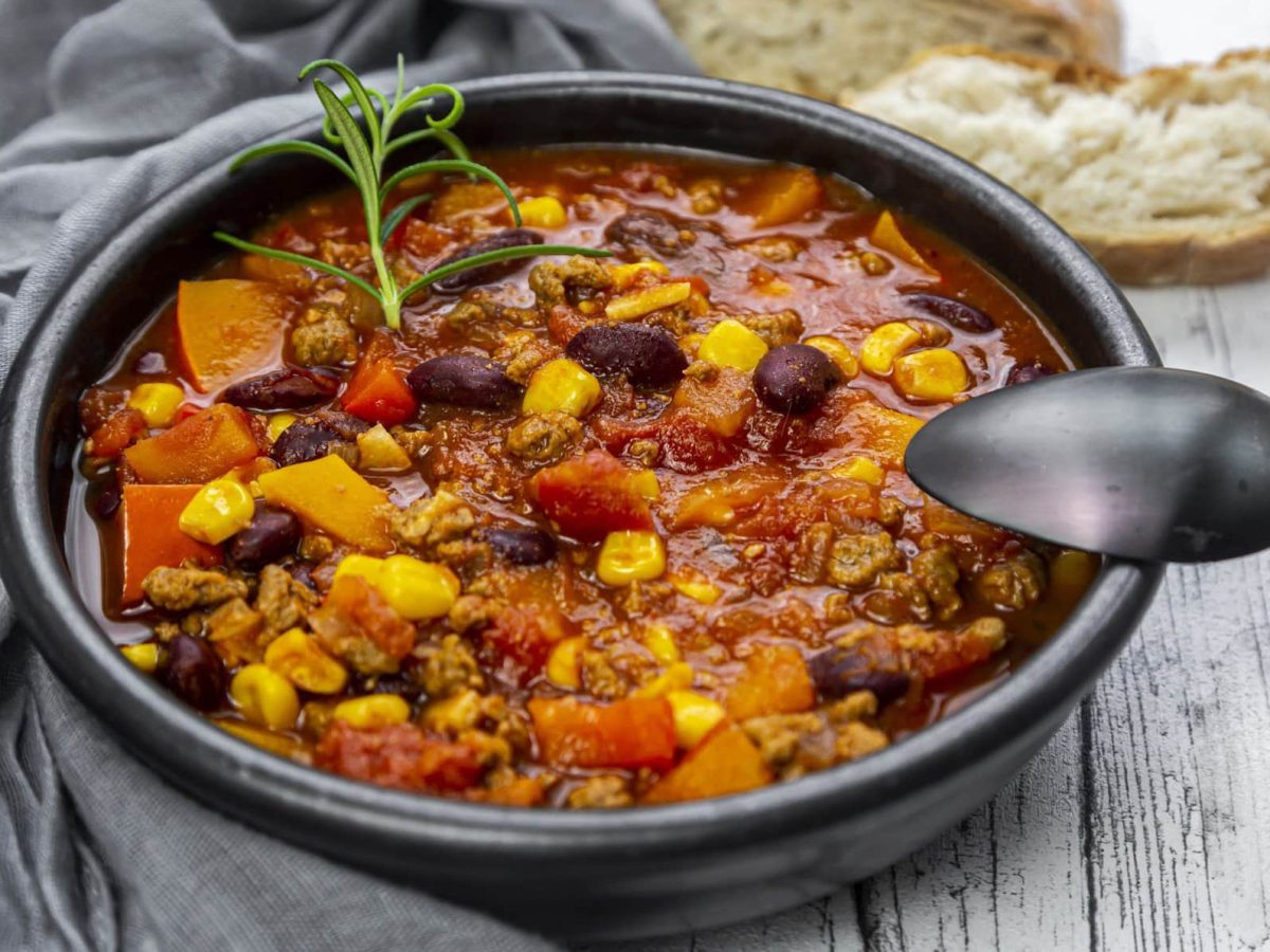 Eine schwarze Schale mit dem Chili con Carne auf einem grauen Holztisch mit grauer Tischdecke auf der linken Seite. Alles von schräg oben fotografiert.