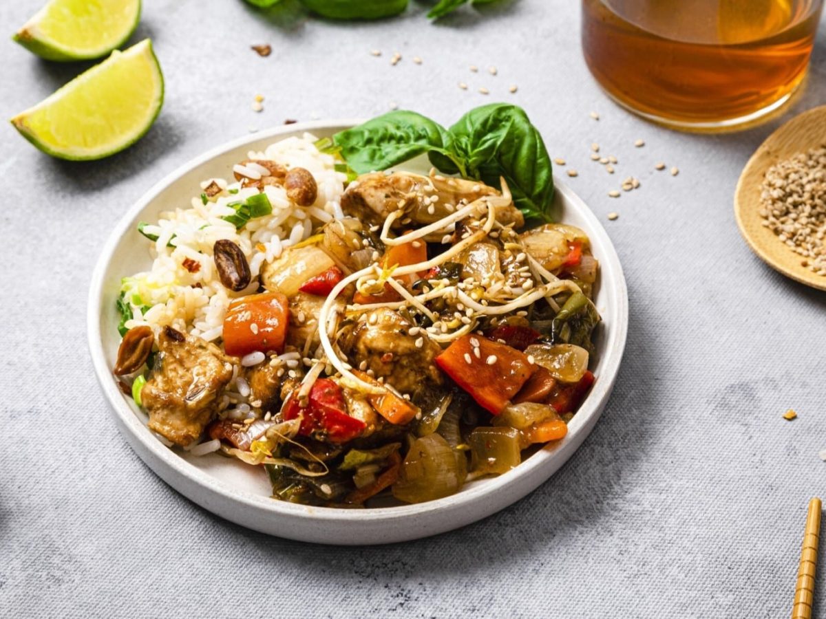 Veganes Chop Suey mit Reis serviert in einem weißen Teller. Rechts Stäbchen, Sesam und Öl. Links davon eine Limette und Chili-Salz. Der Hintergrund ist hellgrau.