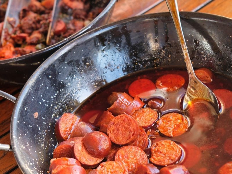 Draufsicht, schräg: ein weiter rustikaler Topf aus Metall mit Chorizos al vino.