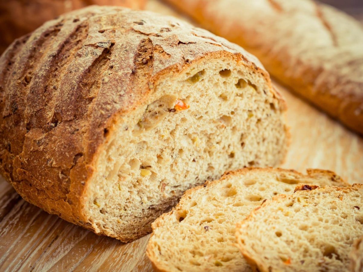 Draufsicht schräg: Ein angeschnittener Laib Ezekiel-Brot liegt auf einem Untergrund aus Holz. Davor liegen einige Scheiben des Brots aus der Bibel.