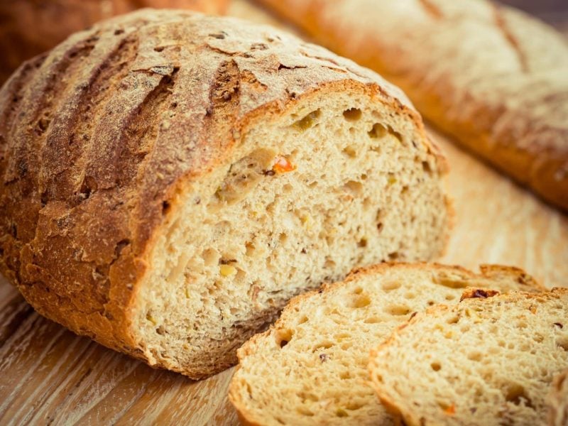 Draufsicht schräg: Ein angeschnittener Laib Ezekiel-Brot liegt auf einem Untergrund aus Holz. Davor liegen einige Scheiben des Brots aus der Bibel.