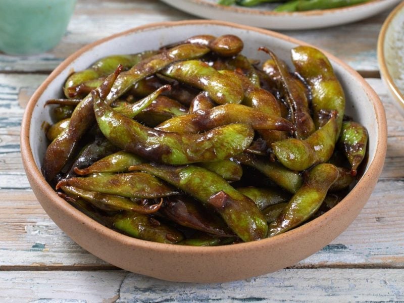 Draufsicht, schräg: Eine Snack-Schale gebratene Edamame mit Ingwer und Sojasauce auf einem hellen Untergrund aus Holz.