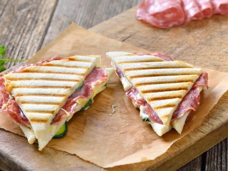 Draufsicht, schräg: Zwei italienische gegrillte Panini mit Salami und Käse liegen auf einem Stück Packpapier, Im Hintergrund ist aufgeschnittene Fenchelsalami.