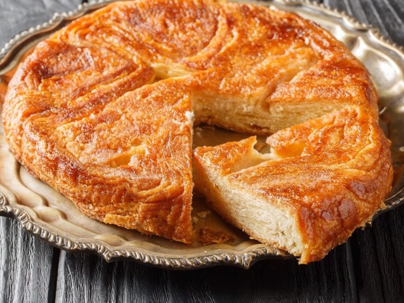 Draufsicht, schräg: Ein Kouign Amann auf einem Platte aus aus silber auf einem Holztisch. Es ist ein Bretonischer Butterkuchen.