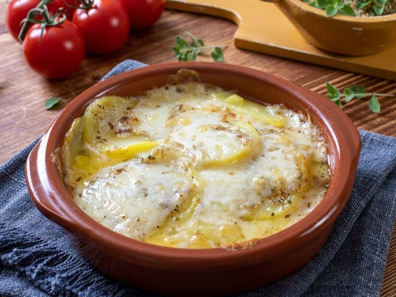 Draufsicht, schräg: Mozzarella-Kartoffeln aus dem Ofen: in einer ofenfestenForm stehen auf einem Holztisch. Drum herum sind Kräuter und Tomaten.