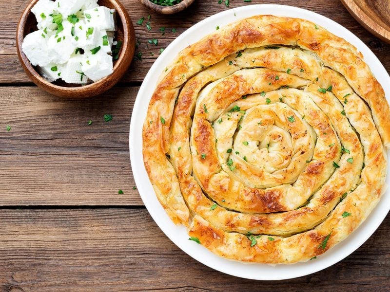 Die Feta-Spinat-Schnecke von oben auf einem dunklen Holztisch fotografiert. Daneben noch ein Schälchen mit Feta und eines mit gehackter Petersilie.