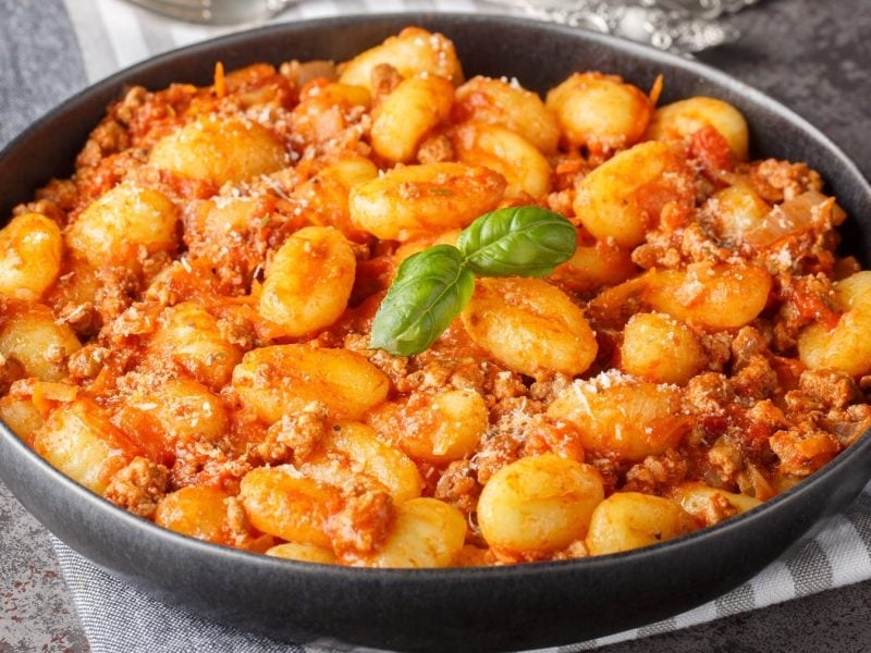 Ein schwarzer Teller mit Gnocchi-Bolognese und einem BAsilikumblatt auf einem grau-weiß-gestreiftem Tuch und grauer Tischplatte. Alles von schräg oben fotografiert.