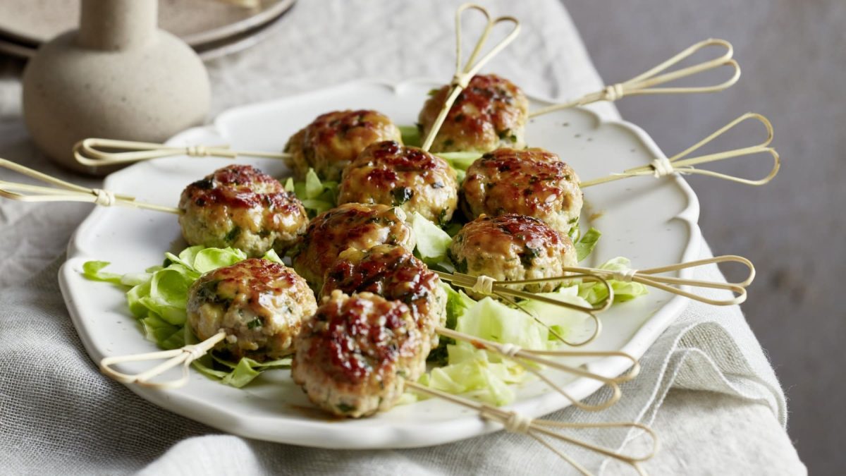 Honigglasierte Mett-Lollies auf einem weißen Servierteller auf einem Tisch mit beigen Tischtuch. Dahinter eine beige Vase und Teller.