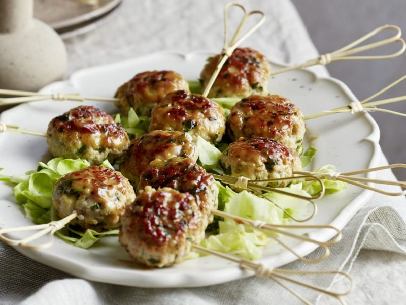 Honigglasierte Mett-Lollies auf einem weißen Servierteller auf einem Tisch mit beigen Tischtuch. Dahinter eine beige Vase und Teller.