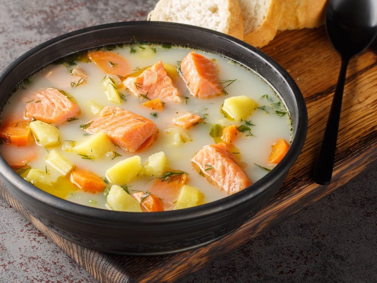 Eine schwarze Schale mit der Kartoffel-Lachs-Suppe auf einem kleinen Holzbrett und grauem Tisch. Alles von schräg oben fotografiert.