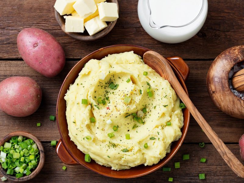 Draufsicht: Untergrund aus Holz. Eine runde Tonschlüssel Kartoffelpüree mit Rettich. Drum herum liegen rotschalige Kartoffeln, Schnittlauchröllchen, Butterwürfel und ein Holzlöffel.