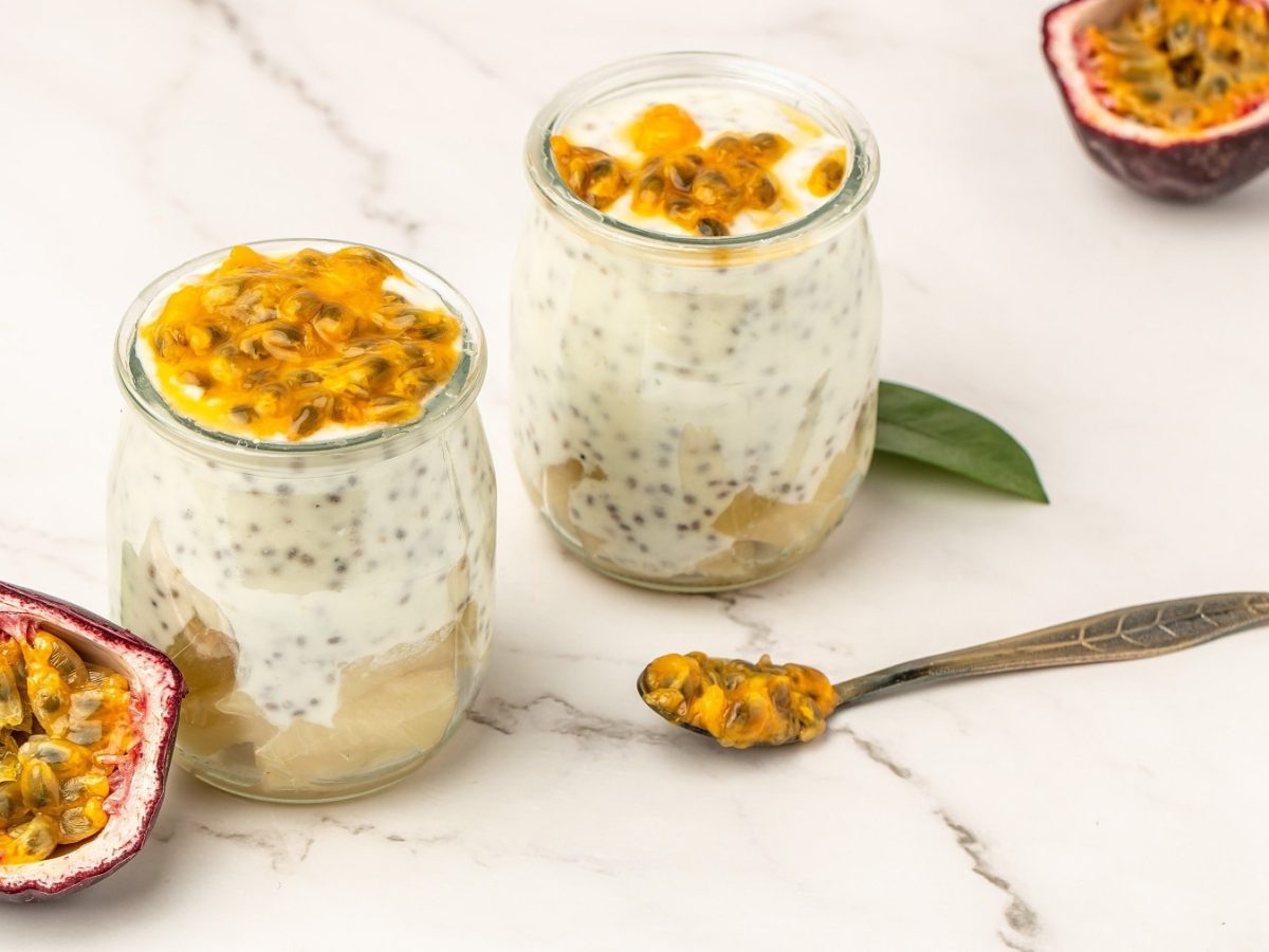 Zwei Gläser mit dem Kokos-Chia-Pudding auf einem hellen Marmortisch. Daneben zwei halbierte Passionsfrüchte und zwei grüne Blätter. Alles von schräg oben fotografiert.