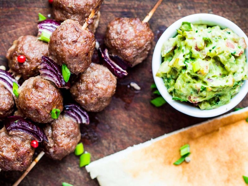 Draufsicht: Drei Spieße Lammbuletten liegen auf einem Untergrund aus Holz. Daneben ist eine Schüssel mit einem Avocado-Dip.