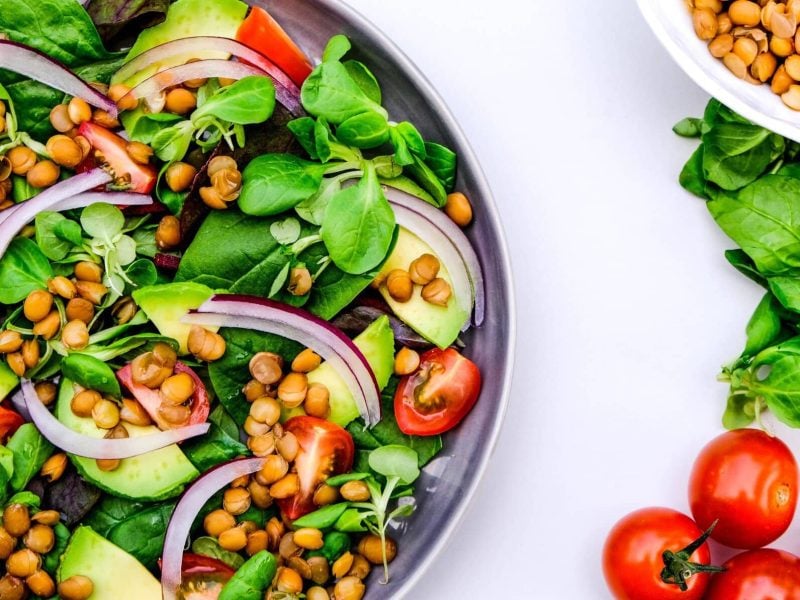 Draufsicht: Ein halbe Schüssel Linsen-Avocado-Salat. Daneben liegen kleine Tomaten, Spinatblätter und Linsen.