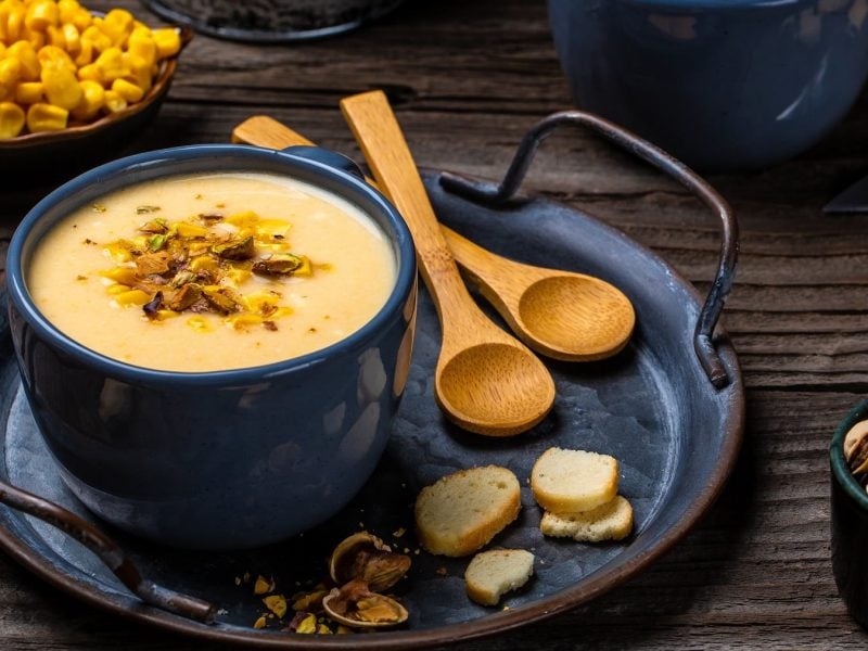 Die Maissuppe in dunkelblauem Geschirr auf einem rustikalen Holztisch. Alles von schräg oben fotografiert.