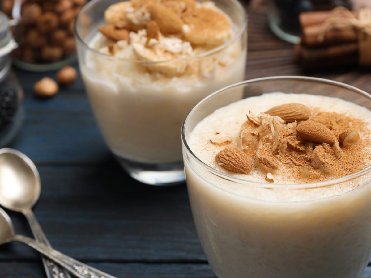 Zwei Gläser mit dem Mandel-Milchreis auf einem dunkelblauen Holztisch mit zwei silbernen Löffeln sowie Gewürzen.