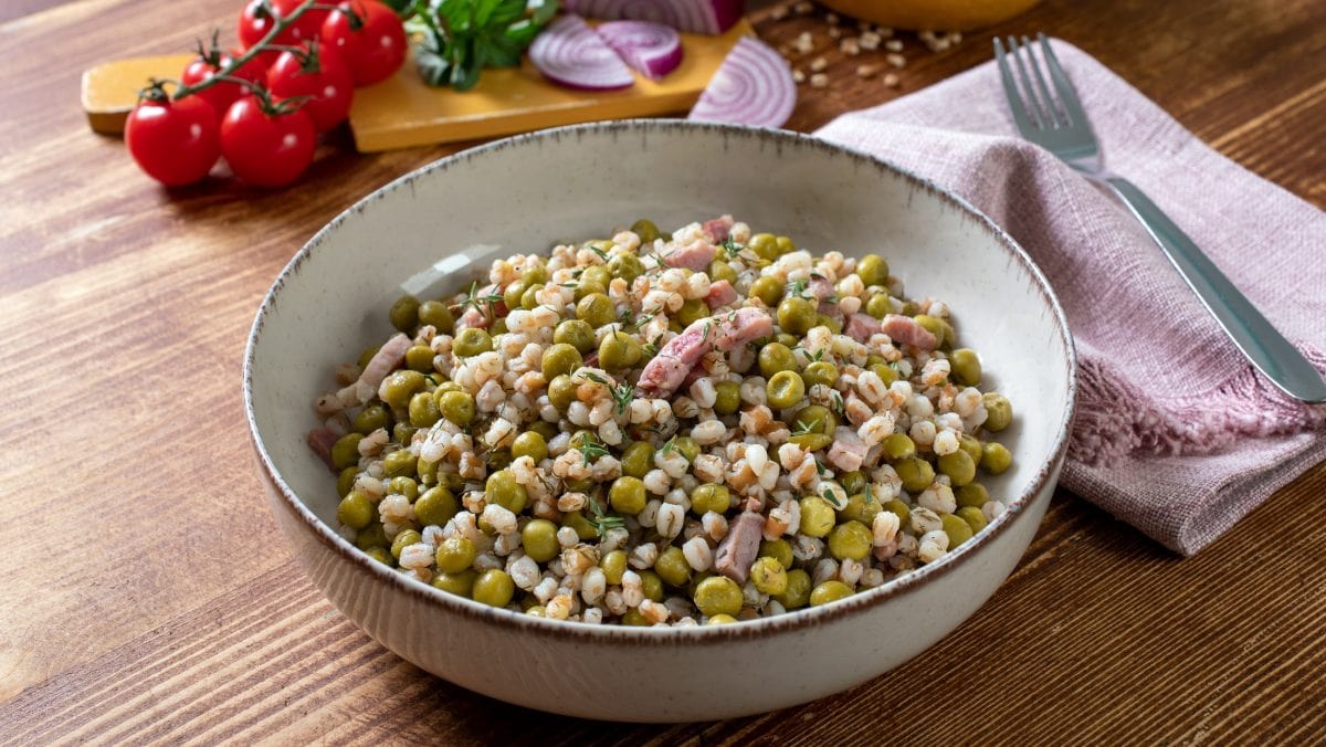 Draufsicht schräg: Eine Schüssel mit einem Perlgraupensalat mit Erbsen und Speck steht auf einem Holztisch.