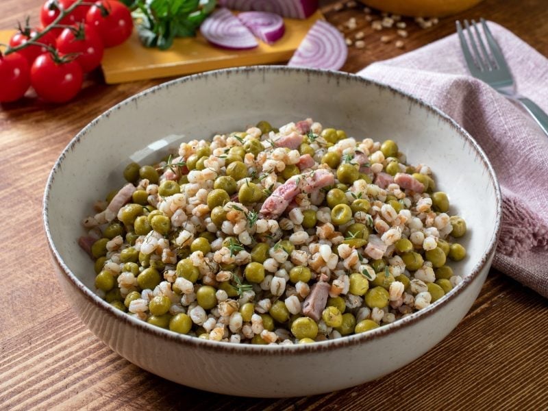 Draufsicht schräg: Eine Schüssel mit einem Perlgraupensalat mit Erbsen und Speck steht auf einem Holztisch.