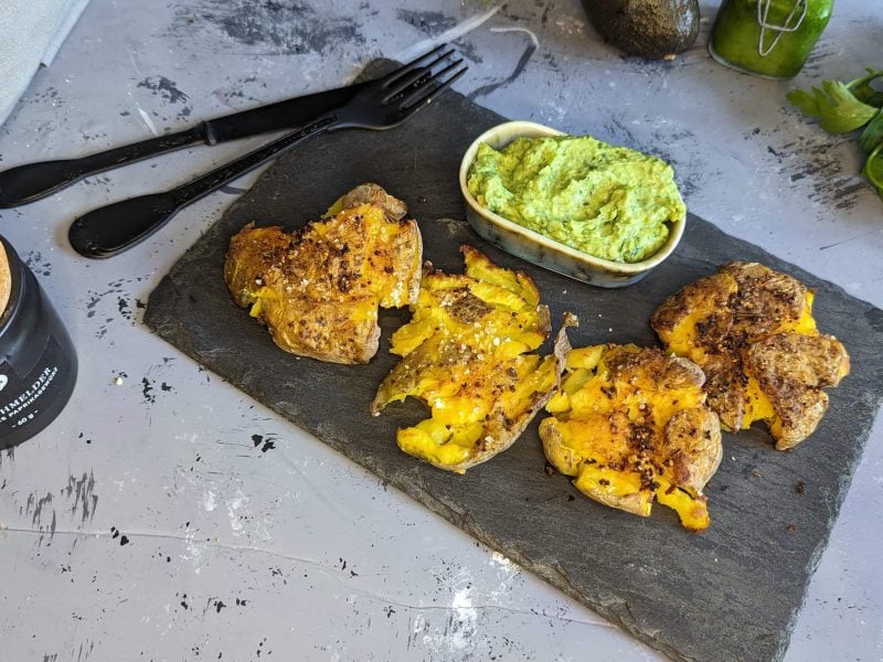 Eine Schieferplatte mit den Kartoffeln und dem Dip auf einem grauen Tisch mit Avocado und Petersilie. Alles von schräg oben fotografiert.