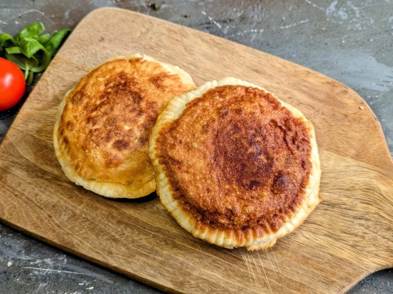 Zwei frittierte Pizzen auf einem Holzbrett und grauem Untergrund. Daneben Tomate und Basilikum. Alles von schräg oben fotografiert.