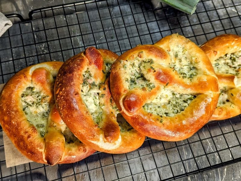 Mehrere Flammkuchen-Brezeln auf einem schwarzen Gitter und grauem Untergrund. Alles direkt von oben fotografiert.